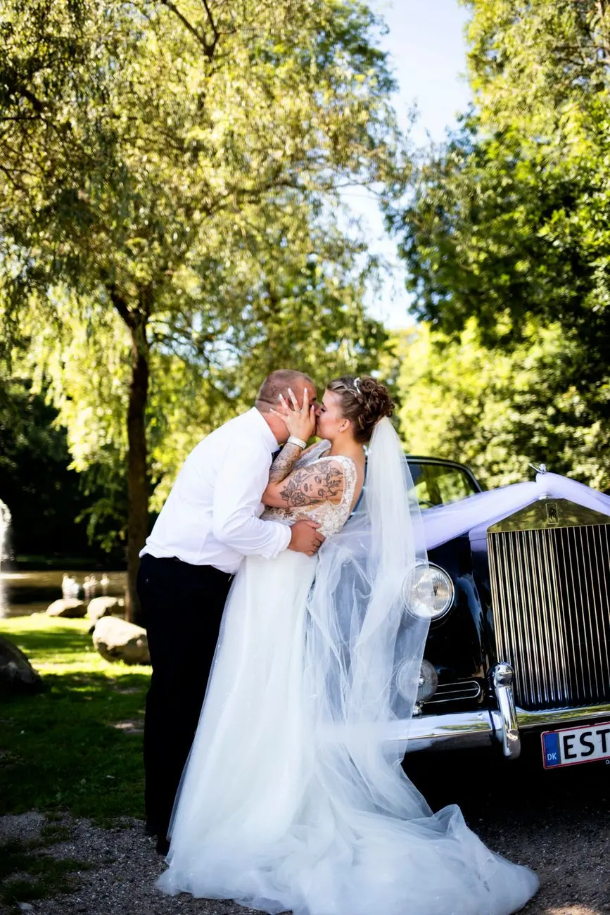 A bride and groom kiss beside a vintage car